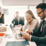 Two cheerful colleagues in formal wear solving problem and looking at paperwork. In background employees talking. It is literally true that succeed best and quickest by helping others to succeed.
