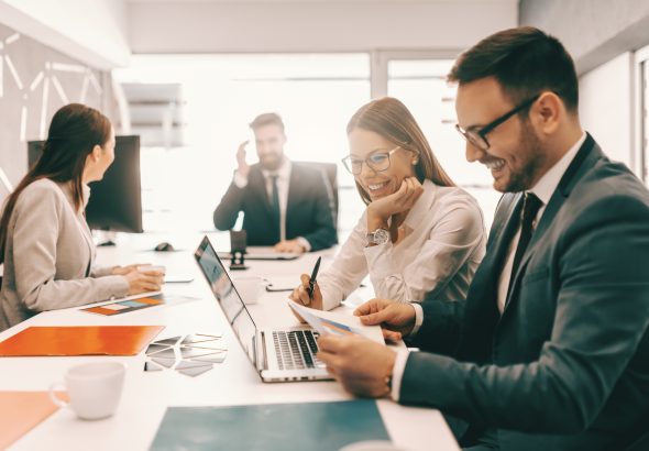 Two cheerful colleagues in formal wear solving problem and looking at paperwork. In background employees talking. It is literally true that succeed best and quickest by helping others to succeed.